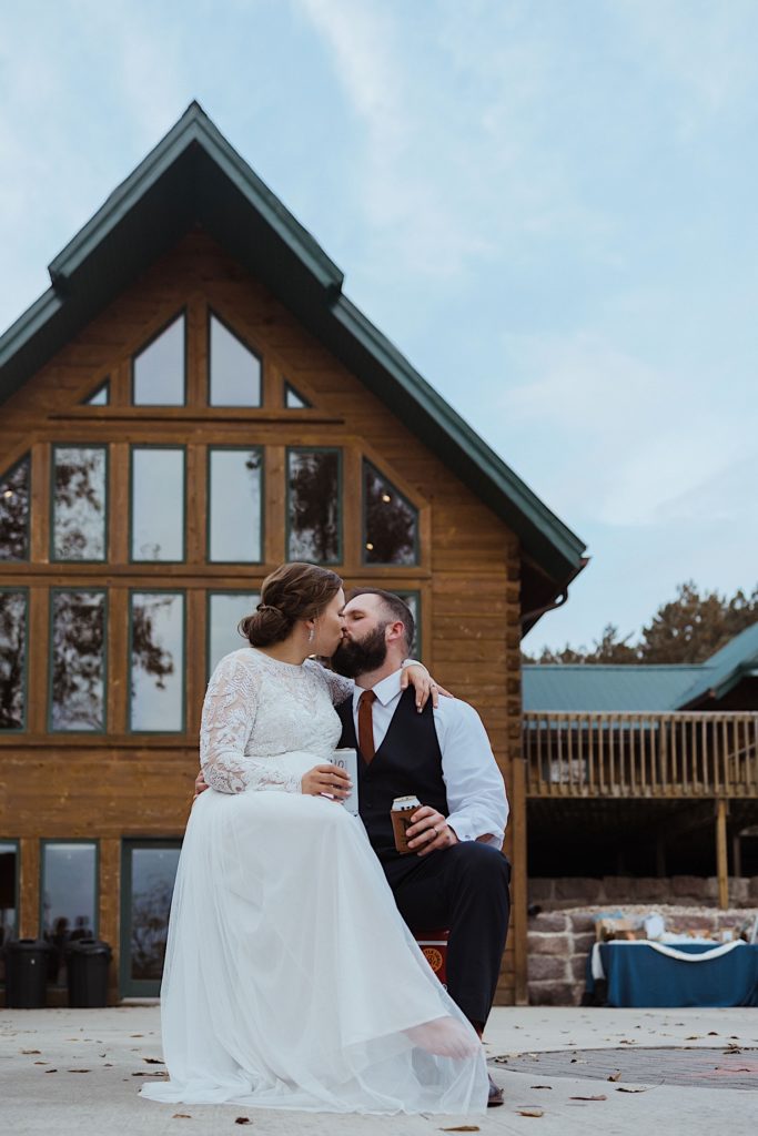 Bride and groom sit and kiss in front of wood cabin holding beers