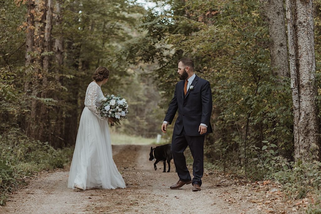 Bride and groom walking through a forest on a dirt road look back at their dog