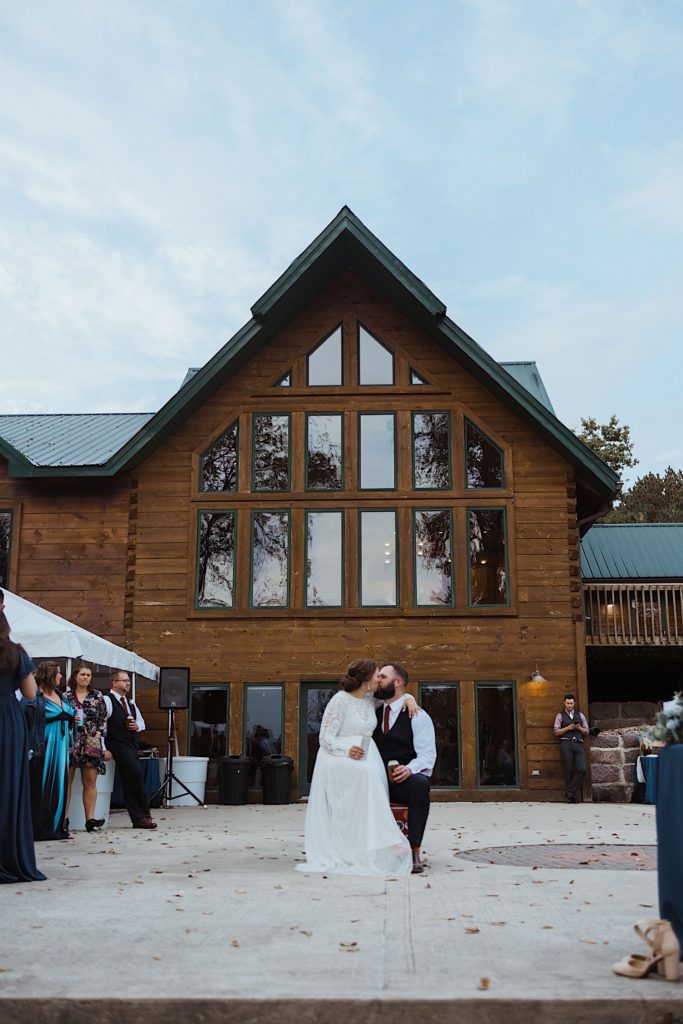 Bride and groom sit and kiss in front of wood cabin holding beers with wedding guests to either side of them