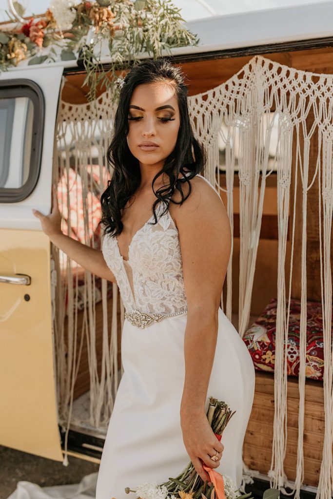 Photo of bride in her wedding dress standing outside of styled van holding flowers