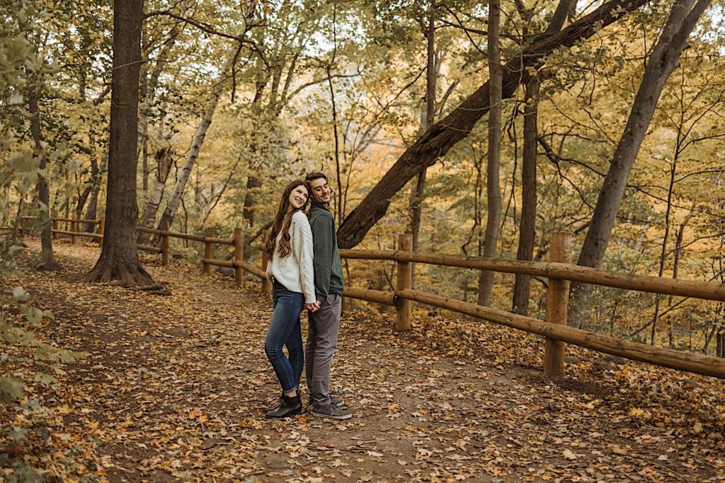 Couple stands back to back next to wood fence along a path in a Wisconsin forest preserve