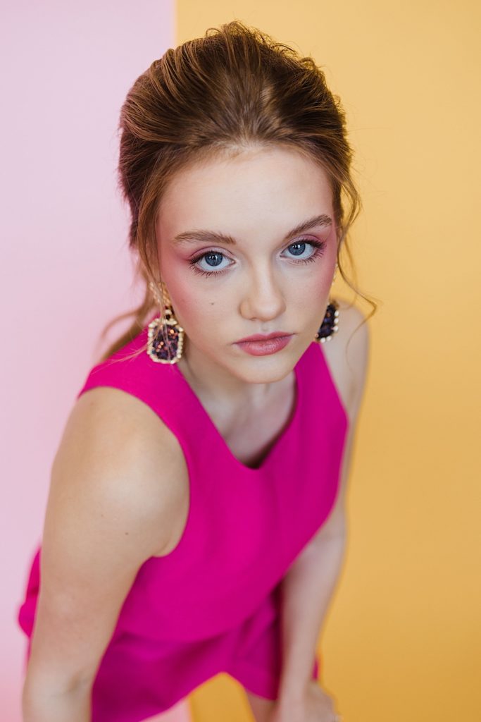 Woman wearing a pink dress, ruby earrings, and pink makeup looks at the camera with a half pink half dark orange background