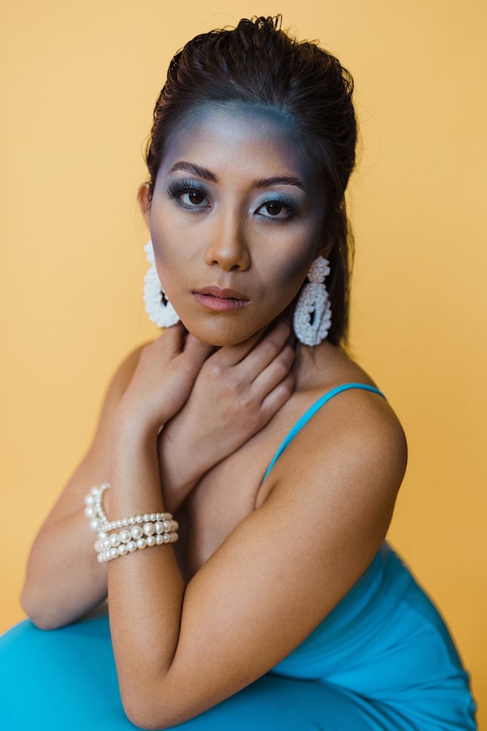 Woman wearing a blue dress, white earrings and bracelets, and blue makeup looks at the camera with a dark yellow background
