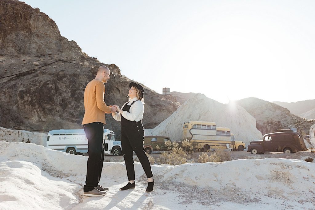 Couple hold hands and look at one another with old busses and cars and mountains in the background