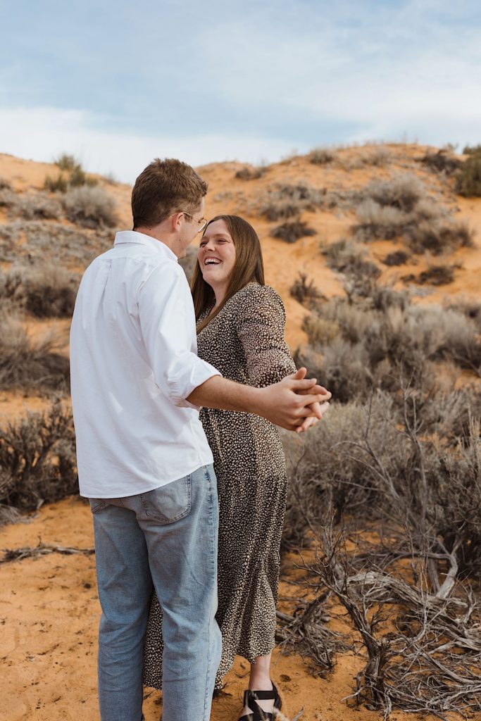 Couple hold hands and extend them towards the camera while smiling at one another in the desert