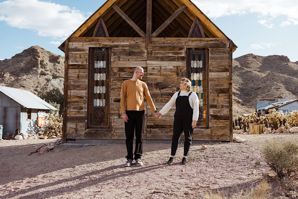 Couple hold hands and look at one another in front of old worn down wood church in the desert