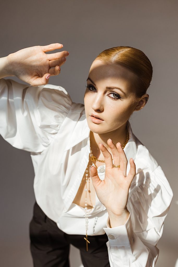 Woman wearing a white blouse, black pants and gold necklace holds her hands up to block the sun while looking at the camera