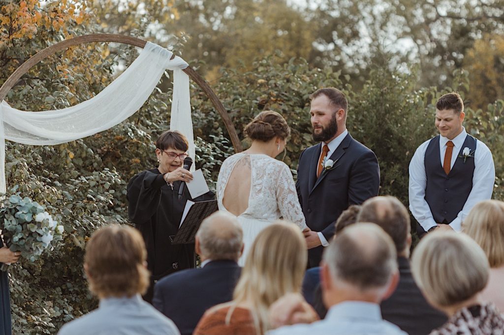 Bride and groom exchanging vows at their wedding ceremony while the guests, groomsmen and pastor watch