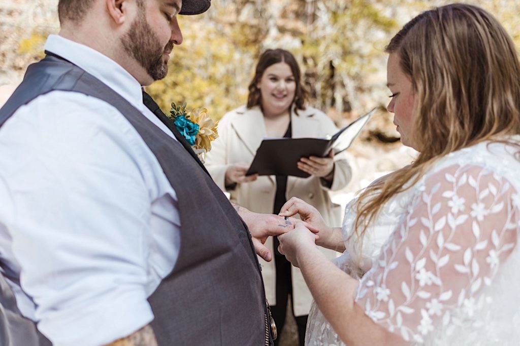 A bride puts a wedding ring on her husband's hand while their officiant stands behind them holding a binder and smiling.