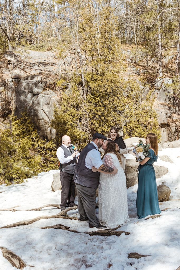 A newly wed couple kiss in front of their two guests and their officiant as they all cheer in a snowy forest