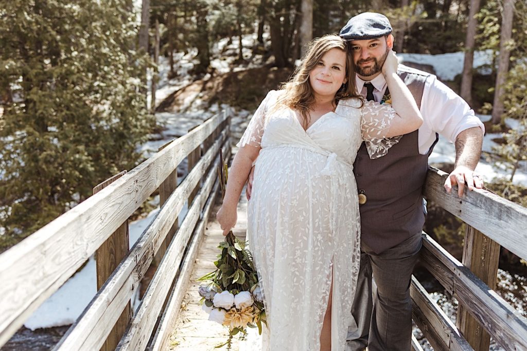 A newly wed couple stand on a wooden bridge in front of a snowy forest. The bride is standing in front of the groom holding a bouquet in one and and resting her other hand on her husbands face as they smile at the camera
