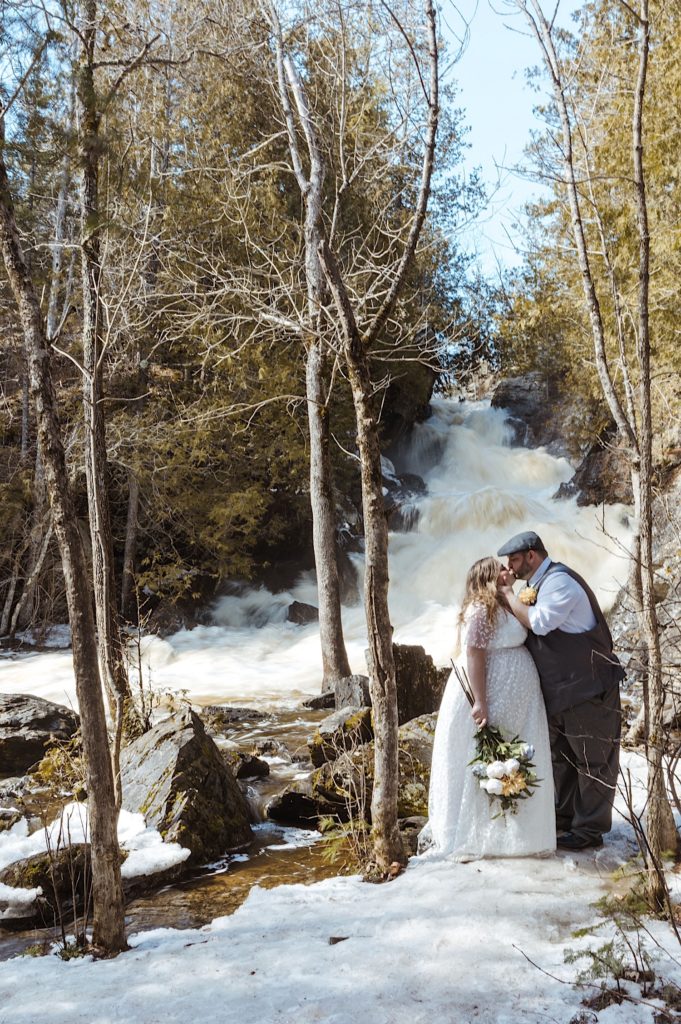 A couple on icy ground in wedding attire kiss next to Longslide Falls in Wisconsin after their elopement.